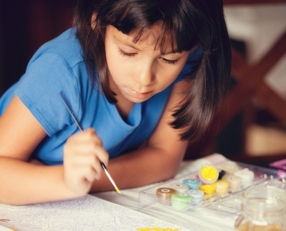 A young child with brown hair and a green shirt smiles and holds a blue object toward the camera in a brightly lit room with a blurred background.